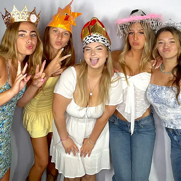 Five young women posing and smiling with silly faces, wearing colorful props in a Glamoor Photo Booth